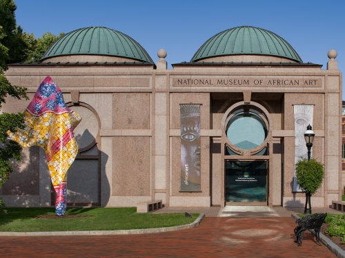Exterior of concrete building with two domes, glass entry, and a colorful, abstract sculpture in front.