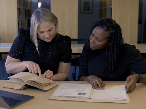 Two women sit side-by-side at a table, looking at archival pages and books in a library setting.